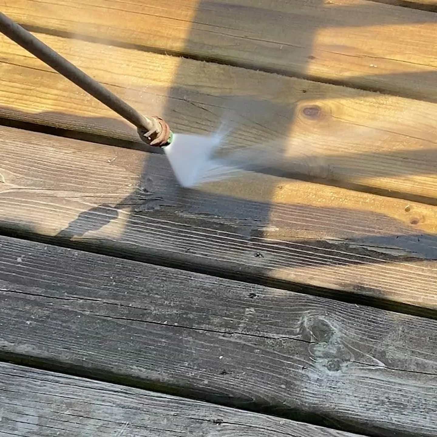 A weathered wood deck being pressure washed, the wand cutting a bright gold stripe through grey boards